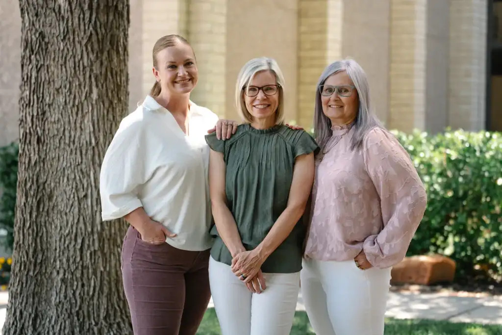 Three women standing together outside, smiling warmly in coordinated outfits—professional team portrait for a healthcare or wellness brand.