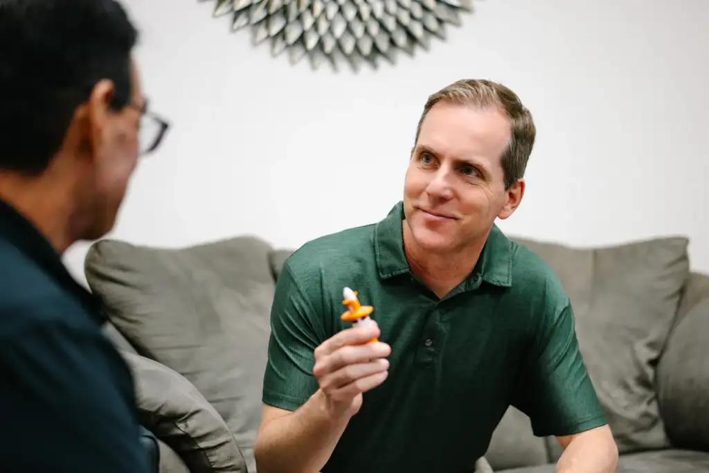A man in a green polo shirt sits on a couch, smiling and holding a demonstration nasal spray device while talking with another person. The setting appears relaxed and conversational, suggesting a patient education or healthcare consultation.