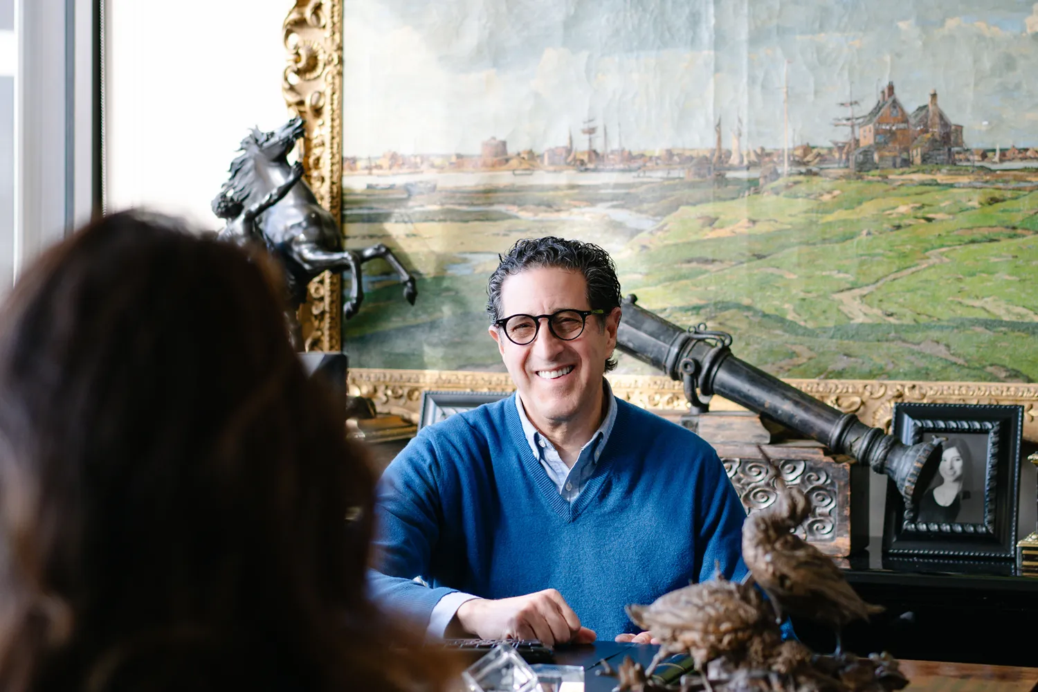 A smiling man in a blue sweater and glasses sits at a desk, engaged in conversation with someone off-camera. Behind him is an ornate gold-framed painting, a horse statue, and a framed photo, creating a warm and personal office environment.