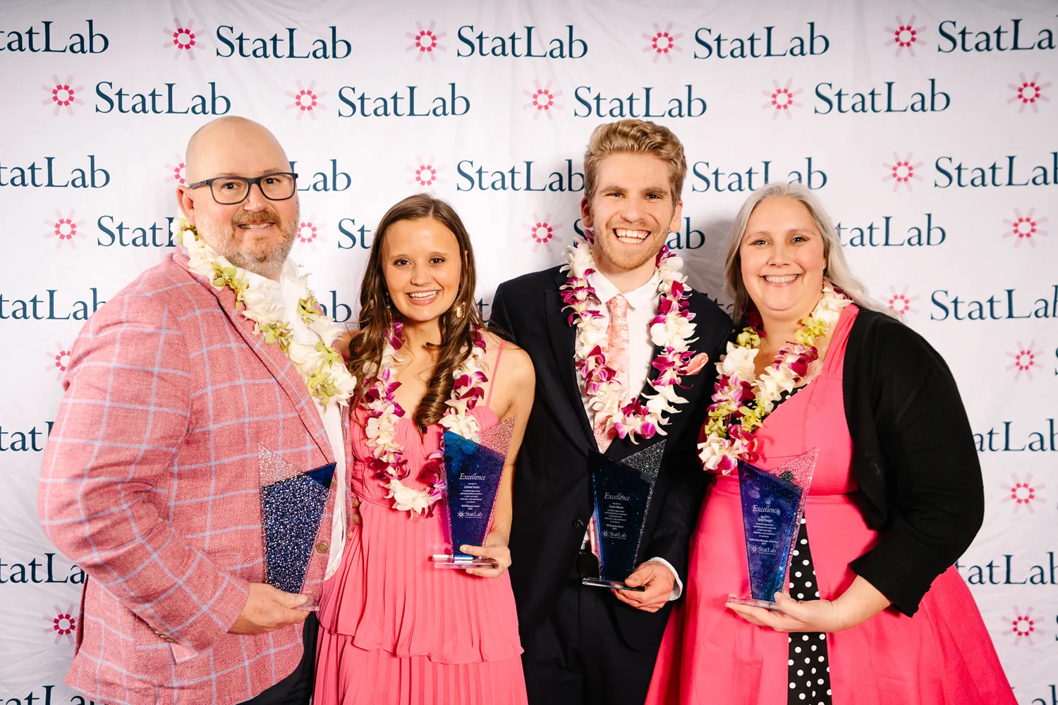 Four award winners wearing pink and black outfits, smiling and holding trophies in front of a StatLab step-and-repeat banner at a Dallas award ceremony.