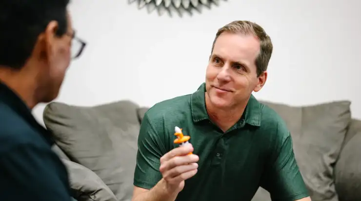 A man in a green polo shirt sits on a couch, smiling and holding a demonstration nasal spray device while talking with another person. The setting appears relaxed and conversational, suggesting a patient education or healthcare consultation.