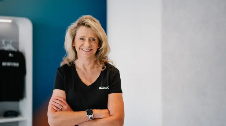 Confident business owner smiling with arms crossed, standing inside a modern fitness studio wearing branded apparel—professional branding headshot with a clean, minimal background.