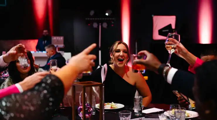 A woman in a black dress laughs as people around her raise glasses for a toast at a StatLab corporate event, with festive pink lighting in the background.