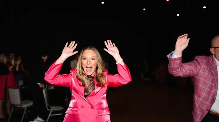 A woman in a bright pink suit raises her hands and smiles enthusiastically on the dance floor at a StatLab event.
