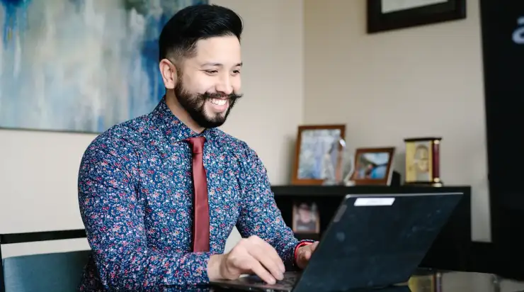 A smiling man in a floral shirt and red tie works on a laptop at his desk in a professional office setting. Framed photos, awards, and a diploma are visible in the background, creating a warm and welcoming atmosphere.
