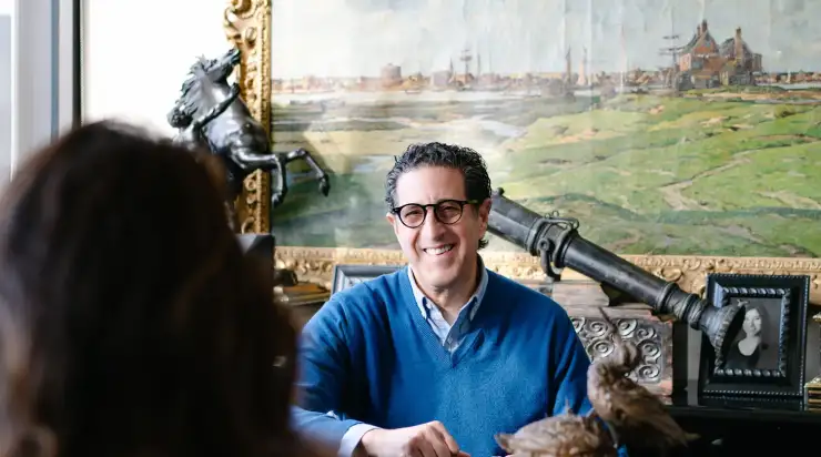 A smiling man in a blue sweater and glasses sits at a desk, engaged in conversation with someone off-camera. Behind him is an ornate gold-framed painting, a horse statue, and a framed photo, creating a warm and personal office environment.