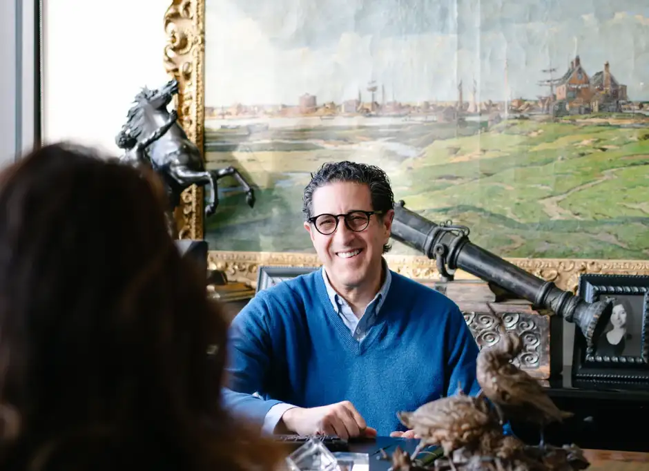 A smiling man in a blue sweater and glasses sits at a desk, engaged in conversation with someone off-camera. Behind him is an ornate gold-framed painting, a horse statue, and a framed photo, creating a warm and personal office environment.