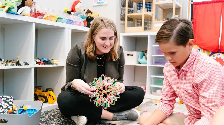 Female therapist smiling while playing with a young client in a colorful, toy-filled therapy room, showcasing a warm and engaging brand atmosphere.
