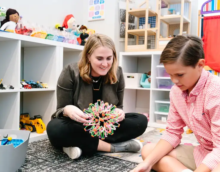 Female therapist smiling while playing with a young client in a colorful, toy-filled therapy room, showcasing a warm and engaging brand atmosphere.
